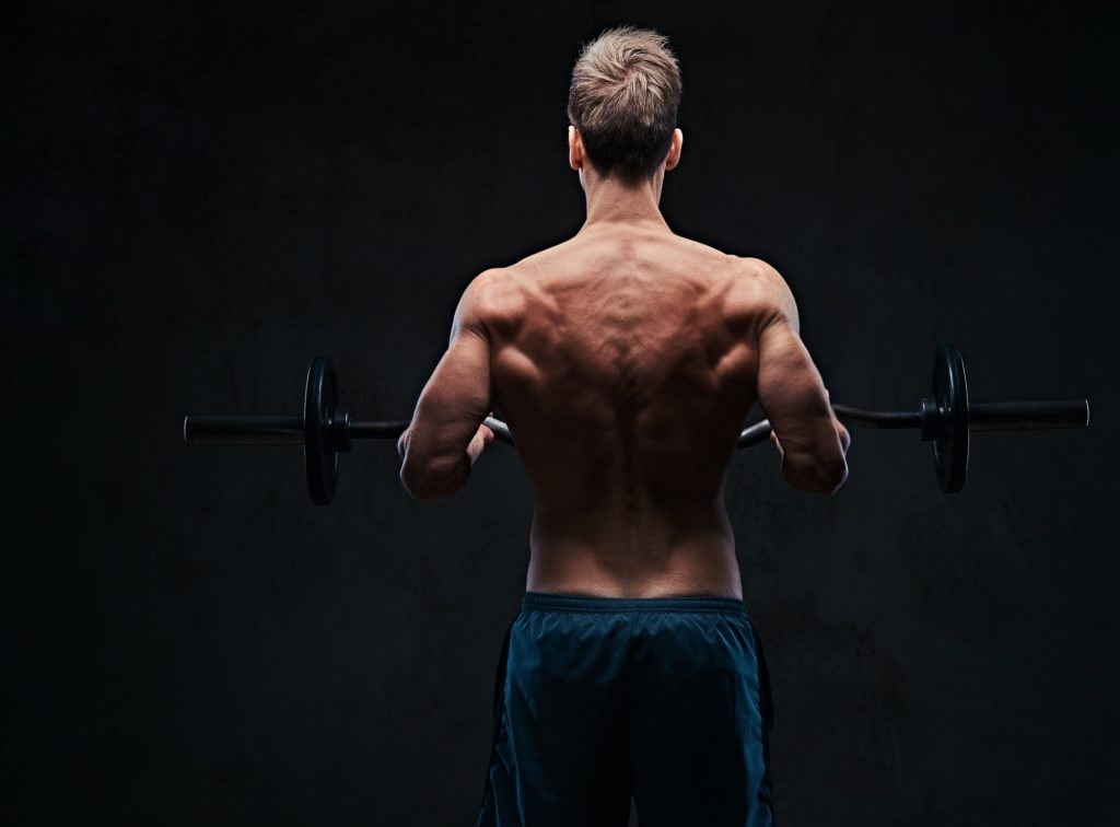 Shirtless male holds barbell over dark grey background.
